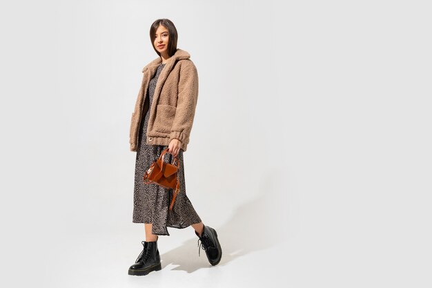 Stylish woman in beige fur coat and dress posing. Full length.