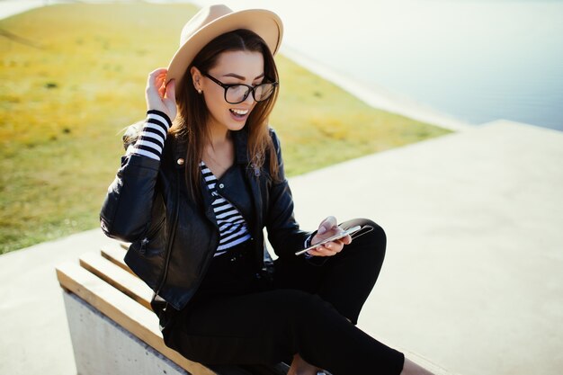 Stylish woman girl sitting in the park near the city lake in cold sunny summer day dressed up in black clothes