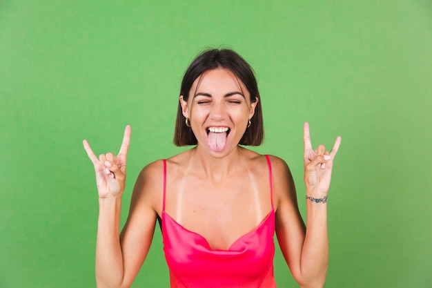 Stylish woman in pink silk dress isolated on green with rock gesture shows her tongue, funny joyful