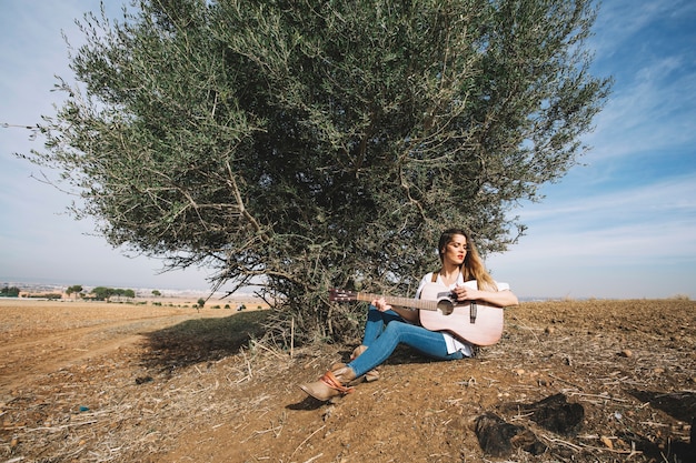 Stylish woman playing guitar near bush