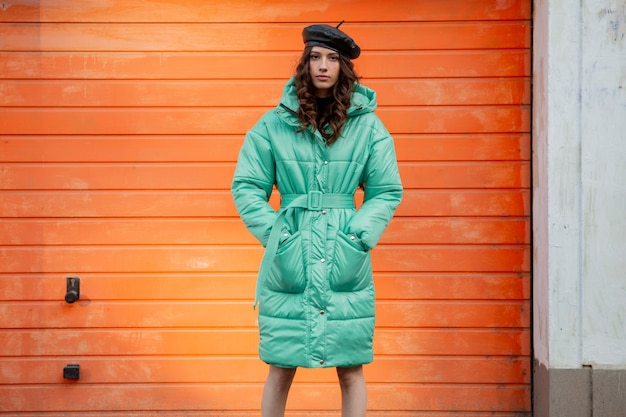 Free Photo stylish woman posing in winter autumn fashion trend puffer coat and hat beret against orange wall in street