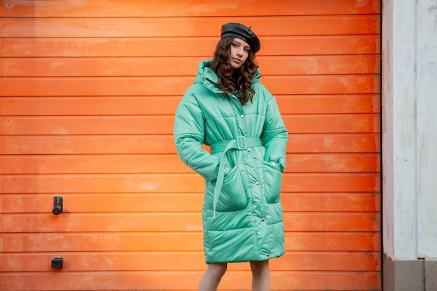 Free photo stylish woman posing in winter autumn fashion trend puffer coat and hat beret against orange wall in street