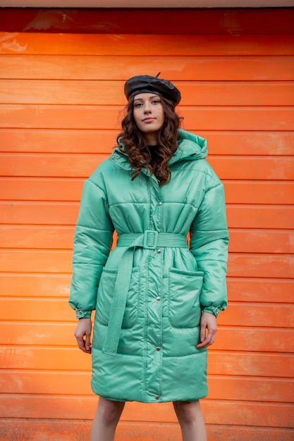 Free Photo stylish woman posing in winter autumn fashion trend puffer coat and hat beret against orange wall in street