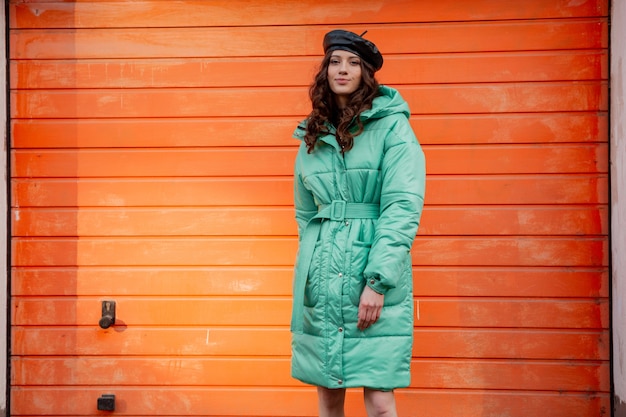 Free Photo stylish woman posing in winter autumn fashion trend puffer coat and hat beret against orange wall in street