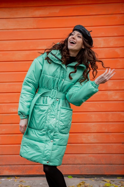 Stylish woman posing in winter autumn fashion trend puffer coat and hat beret against orange wall in street