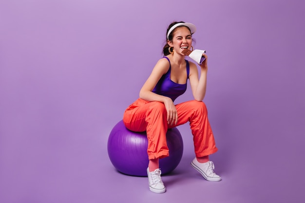 Free photo stylish woman in purple top and red sweatpants eats bar of chocolate sitting on fitball against purple wall