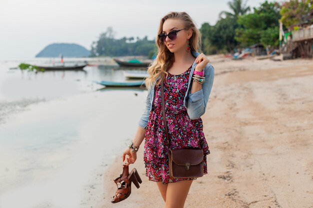 Stylish woman in summer dress vacation walking on beach with shoes in hand