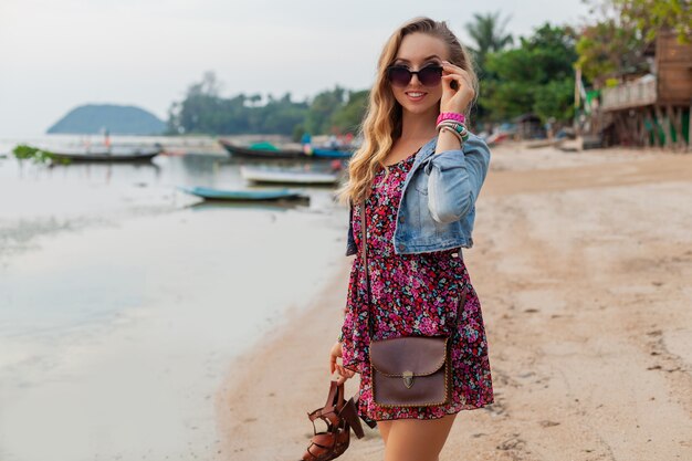 Stylish woman in summer dress vacation walking on beach with shoes in hand