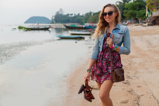 Stylish woman in summer dress vacation walking on beach with shoes in hand