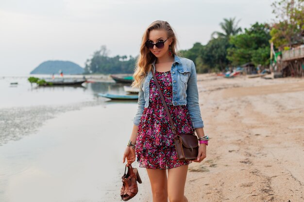 Stylish woman in summer dress vacation walking on beach with shoes in hand