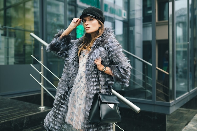 Free Photo stylish woman walking in city in warm fur coat, winter season, cold weather, wearing black cap, holding leather bag, street fashion trend, urban look