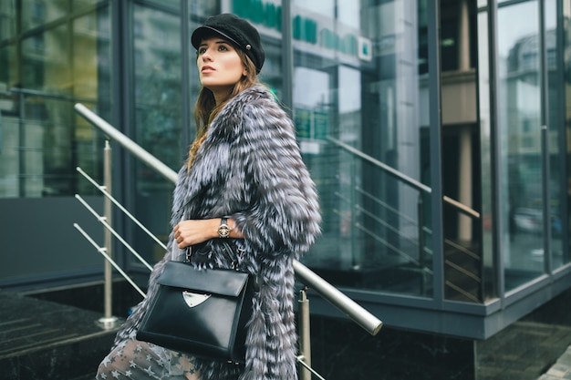 Free photo stylish woman walking in city in warm fur coat, winter season, cold weather, wearing black cap, holding leather bag, street fashion trend, urban look