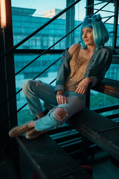 Stylish woman wearing a blue wig sitting on stairs in a modern