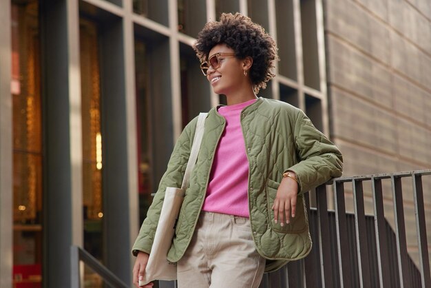 Stylish woman with curly hair wears trendy sunglasses jacket and trousers carries fabric bag has glad expression poses outdoors near urban building spends free time in city People and style concept