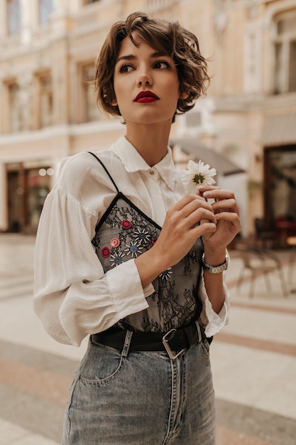 Stylish woman with short hair in jeans with belt holding flower at street. Woman in white blouse with black lace posing in city.