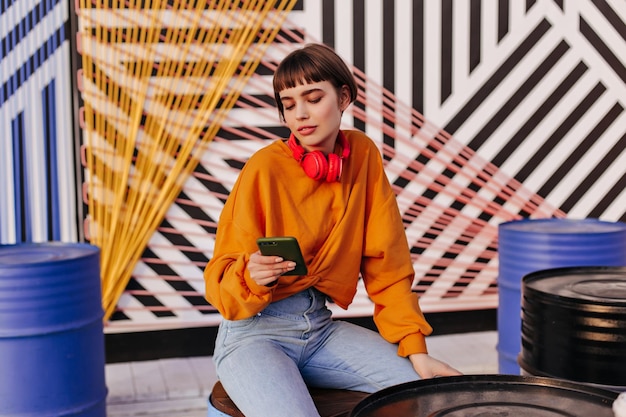 Free photo stylish woman with short hair in orange outfit sitting outdoors charming girl in red headphones and jeans holding phone on striped backdrop