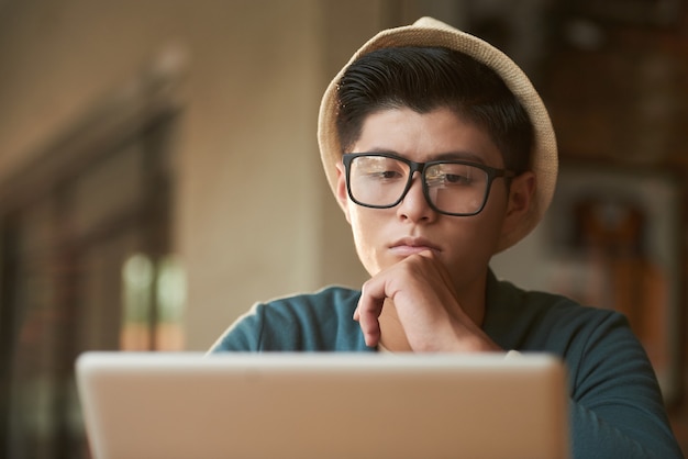 Stylish young Asian man in hat and glasses sitting in cafe and looking at screen