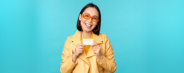 Stylish young asian woman in sunglasses showing credit card and smiling recommending bank contactless payment or discounts in store standing over blue background