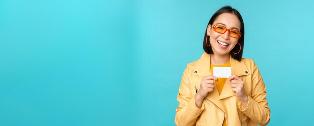 Stylish young asian woman in sunglasses showing credit card and smiling recommending bank contactless payment or discounts in store standing over blue background