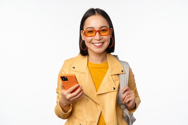 Stylish young asian woman tourist traveller with backpack and smartphone smiling at camera posing against white background