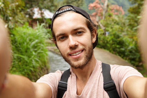 Stylish young bearded male blogger with backpack posing outdoors while recording video or taking selfie