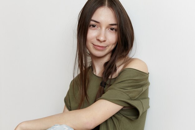 Stylish young Caucasian female with messy hairstyle wearing oversize army green top, looking with happy and joyful smile, spending her day-off indoors. People and lifestyle concept