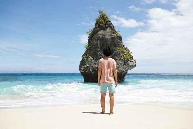 Stylish young Caucasian male adventurer with bare feet standing in front of deserted stone island with vegetation that he finally found, contemplating wonderful seascape, can't believe his eyes