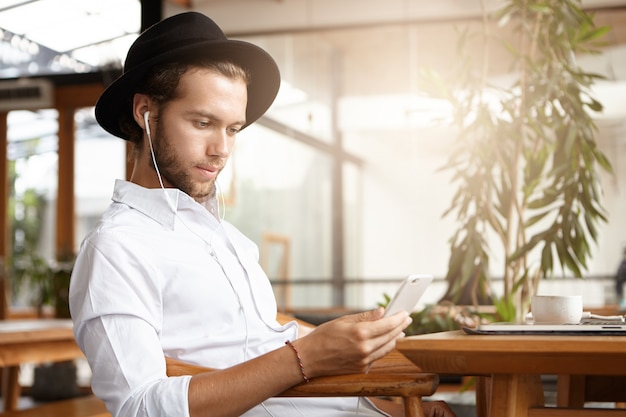 Free Photo stylish young caucasian man in black hat texting sms or reading post via social networks using free wifi on his mobile phone during breakfast at cozy cafe and listening to music on earphones