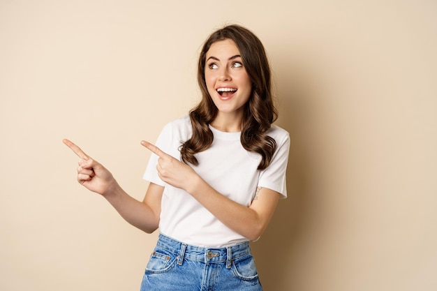 Stylish young caucasian woman smiling, pointing fingers left, showing advertisement, promo offer, standing against beige background.