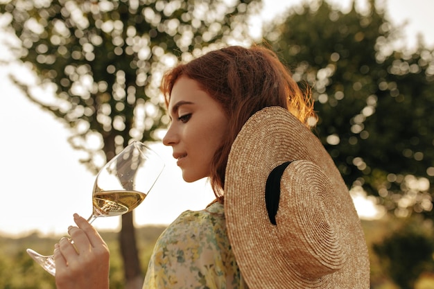 Stylish young girl with foxy hairstyle in modern straw hat and cool summer outfit posing with glass with champagne outdoor