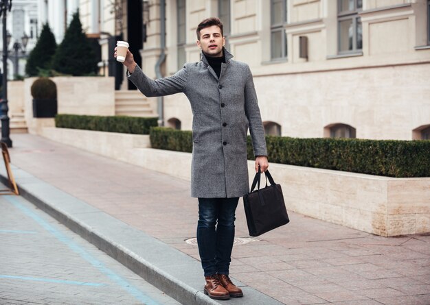 Stylish young man holding takeaway coffee in paper cup, and catching cab on empty street