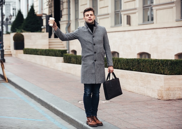 Stylish young man holding takeaway coffee in paper cup, and catching cab on empty street