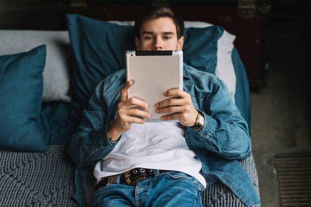 Stylish young man lying on bed looking at digital tablet