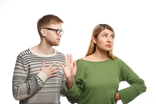 Stylish young man in spectacles holding hand on his chest, asking his angry girlfriend for forgiveness while woman not looking at him, making stop gesture as if saying: I don't need your excuse