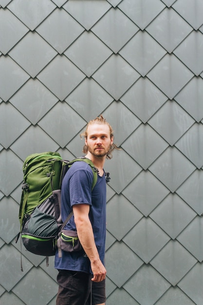 Free photo stylish young man with travelling backpack standing against wall looking away