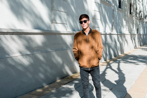 Stylish young teenage boy wearing brown jacket and black sunglasses walking on sidewalk