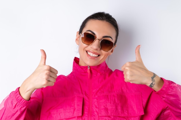 Free Photo stylish young woman in a bright pink sports jacket trendy sunglasses on a white background shows thumbs up with a smile