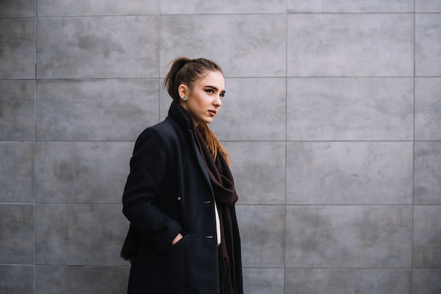 Free Photo stylish young woman in coat with scarf near grey wall