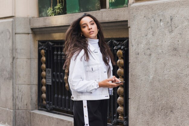 Stylish young woman holding mobile phone in hand looking at camera