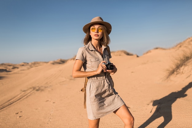 Stylish young woman in khaki dress walking in desert, traveling in Africa on safari, wearing hat and backpack, taking photo on vintage camera