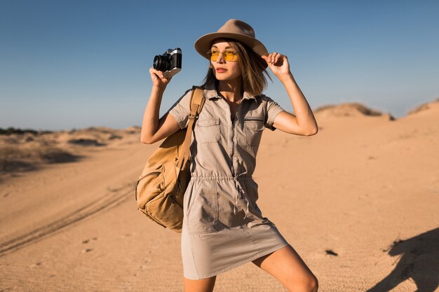 Stylish young woman in khaki dress walking in desert, traveling in Africa on safari, wearing hat and backpack, taking photo on vintage camera