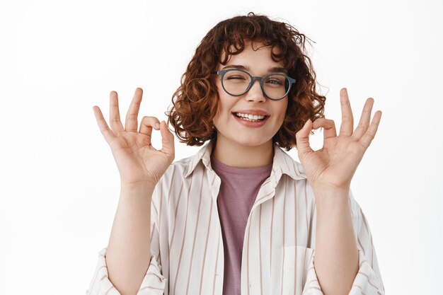 Stylish young woman praise something good, showing okay signs OK and winking, nod in approval, pleased by great quality, make compliment, standing over white background.