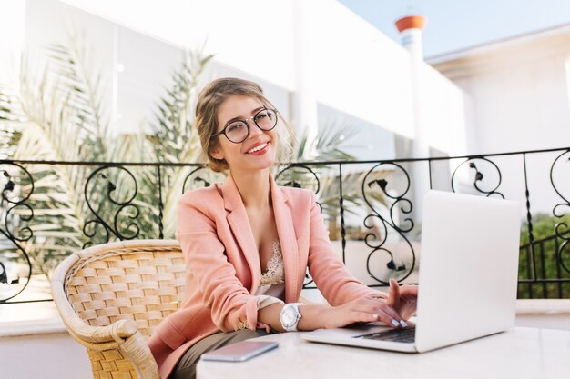 Stylish young woman smiling and working on laptop, joyful girl typing on keyboards, sitting in wicker chair in cozy outdoor cafe. Wearing stylish glasses, pink jacket, beige  blouse, white watches.