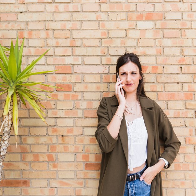 Stylish young woman talking on mobile phone standing against brick wall