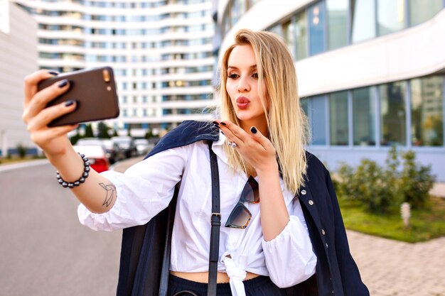 Free Photo stylish young woman wearing trendy navy suit, posing near modern buildings, fashionable accessories , making selfie and sending air kiss to you, positive mood.