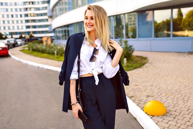 Free photo stylish young woman wearing trendy navy suit, posing near modern buildings, fashionable accessories , smiling end enjoying free sunny summer day, walking near office.