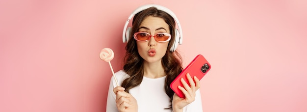 Free Photo stylish young woman with lolipop and cellphone wearing sunglasses and headphones listening music sta