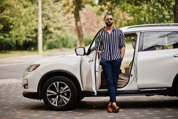 Successful arab man wear in striped shirt and sunglasses pose near his white suv car Stylish arabian men in transport