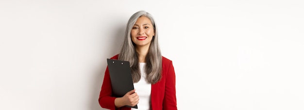 Free Photo successful asian senior business woman holding clipboard wearing red blazer and lipstick at work smi