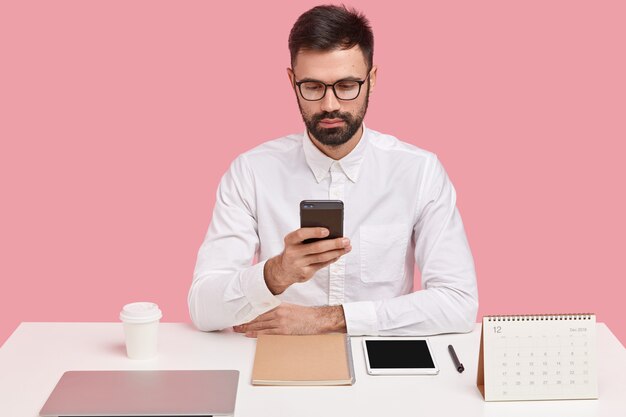 Successful bearded boss in formal white shirt, holds mobile phone, dials telephone number, searches information in browser, being perfectionist